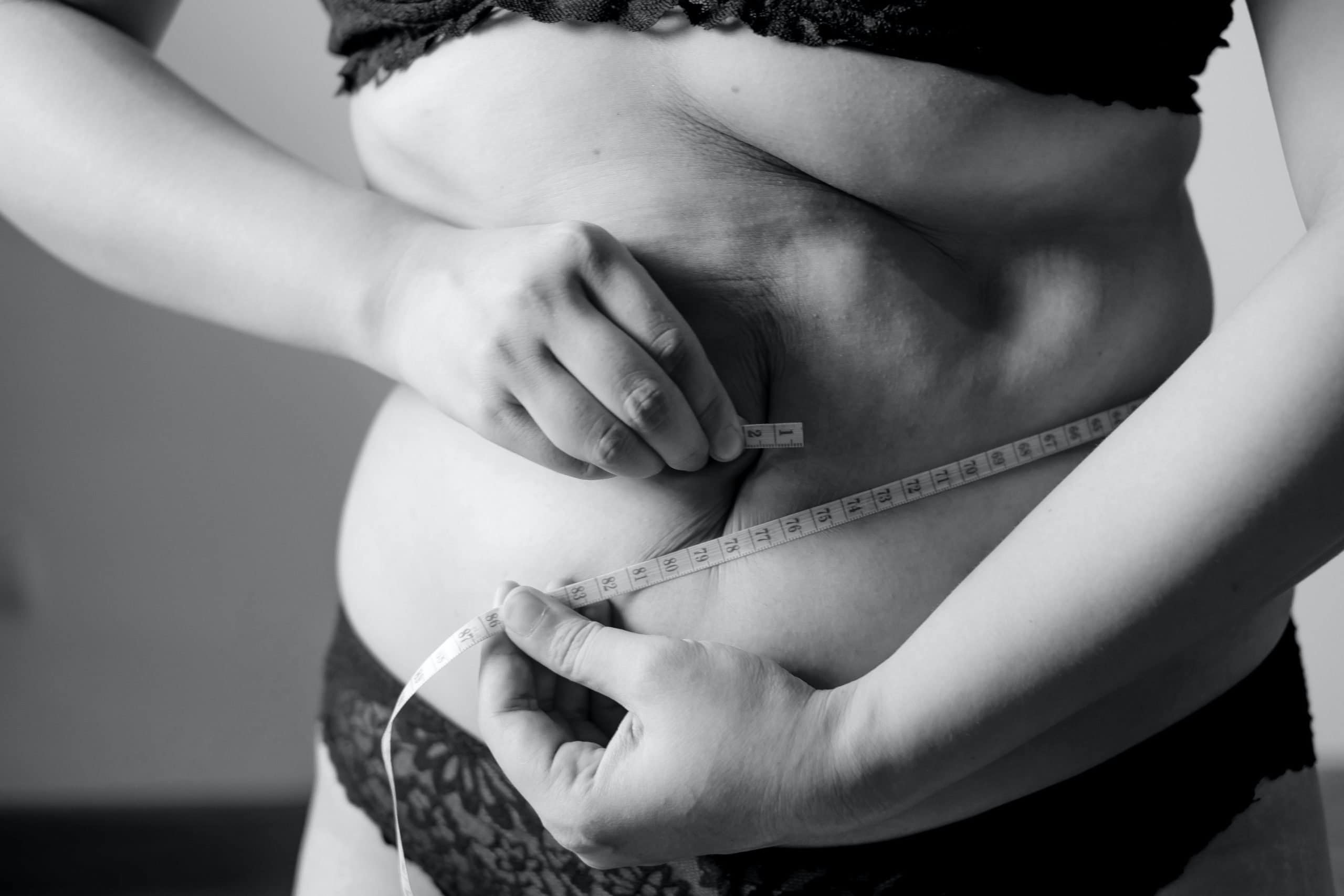 A black and white image of a person measuring their stomach with a measuring tape
